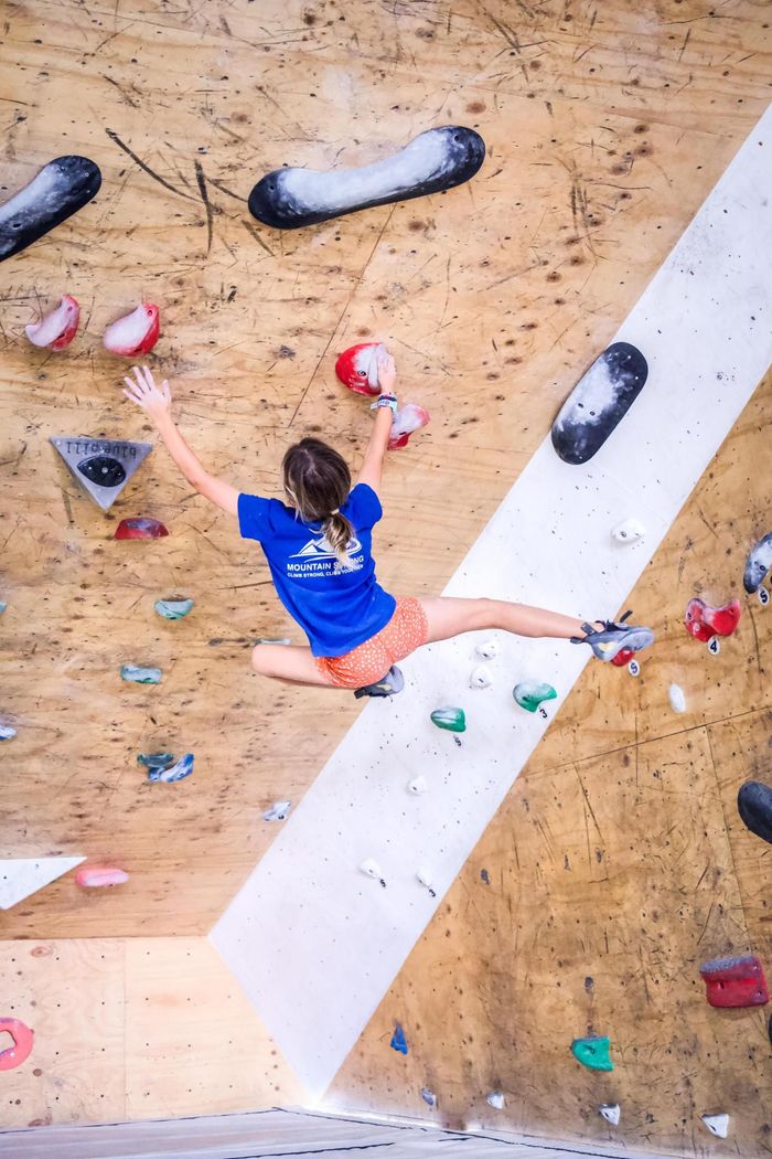Person rock climbing on an angled wall, legs split. Wearing blue shirt and orange shorts, arms outstretched.
