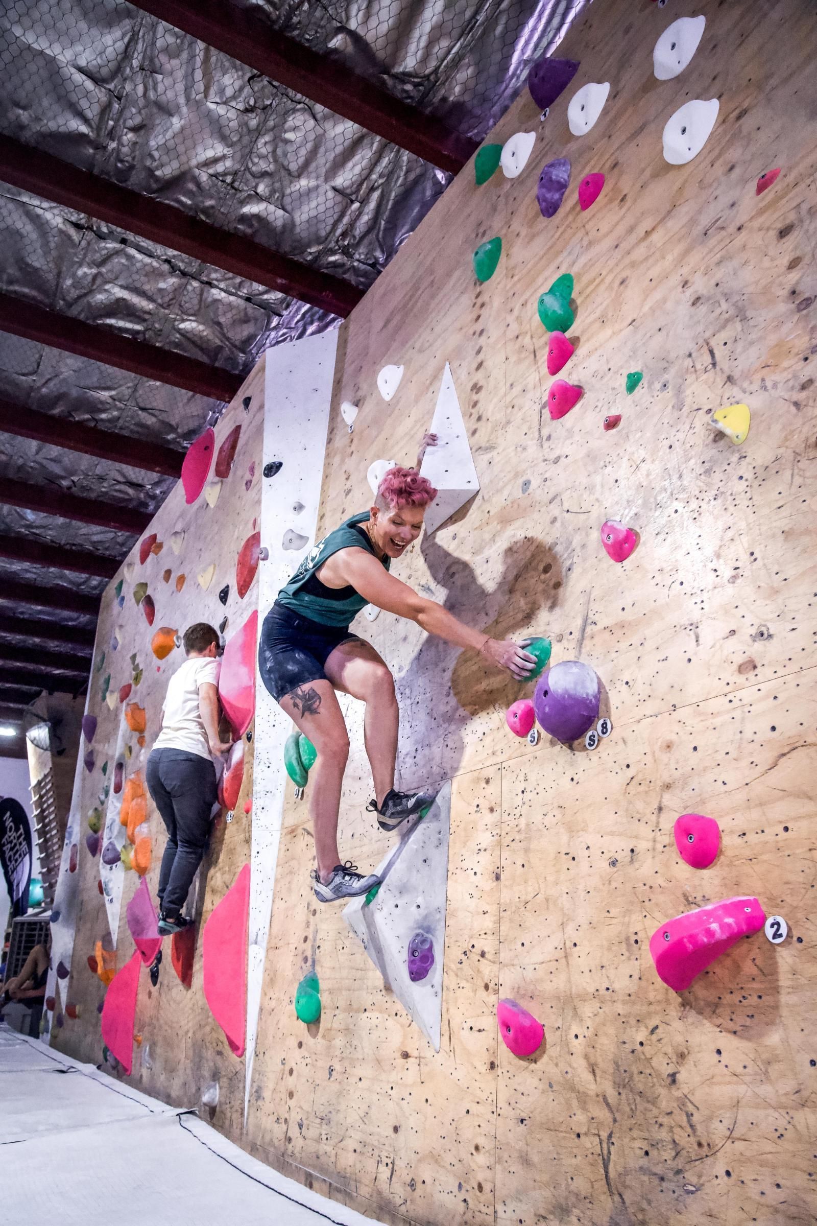 Two people bouldering on a climbing wall with colorful holds. One reaches for a purple hold.
