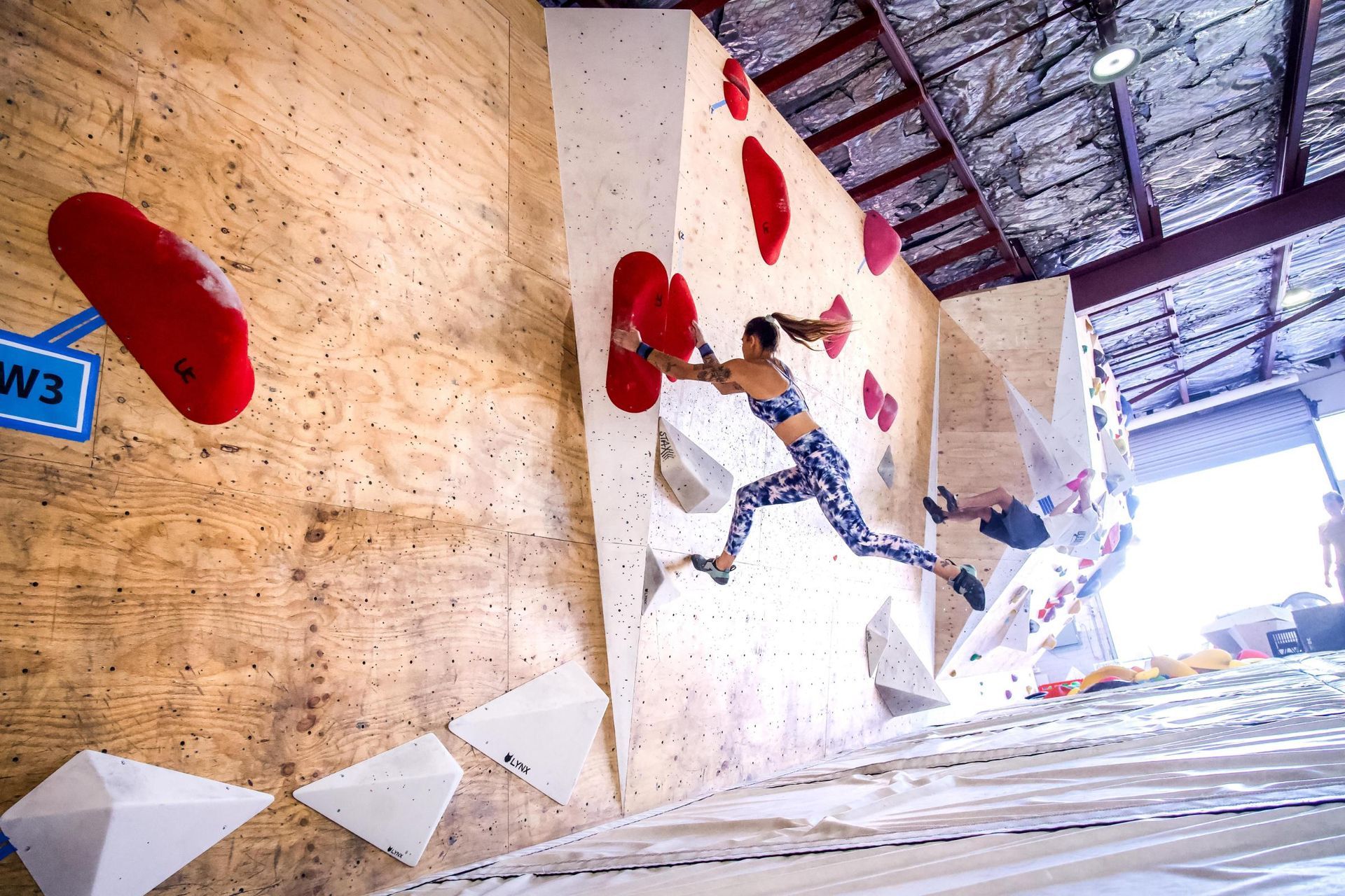 Woman bouldering indoors, jumping for a red hold on a climbing wall with others.