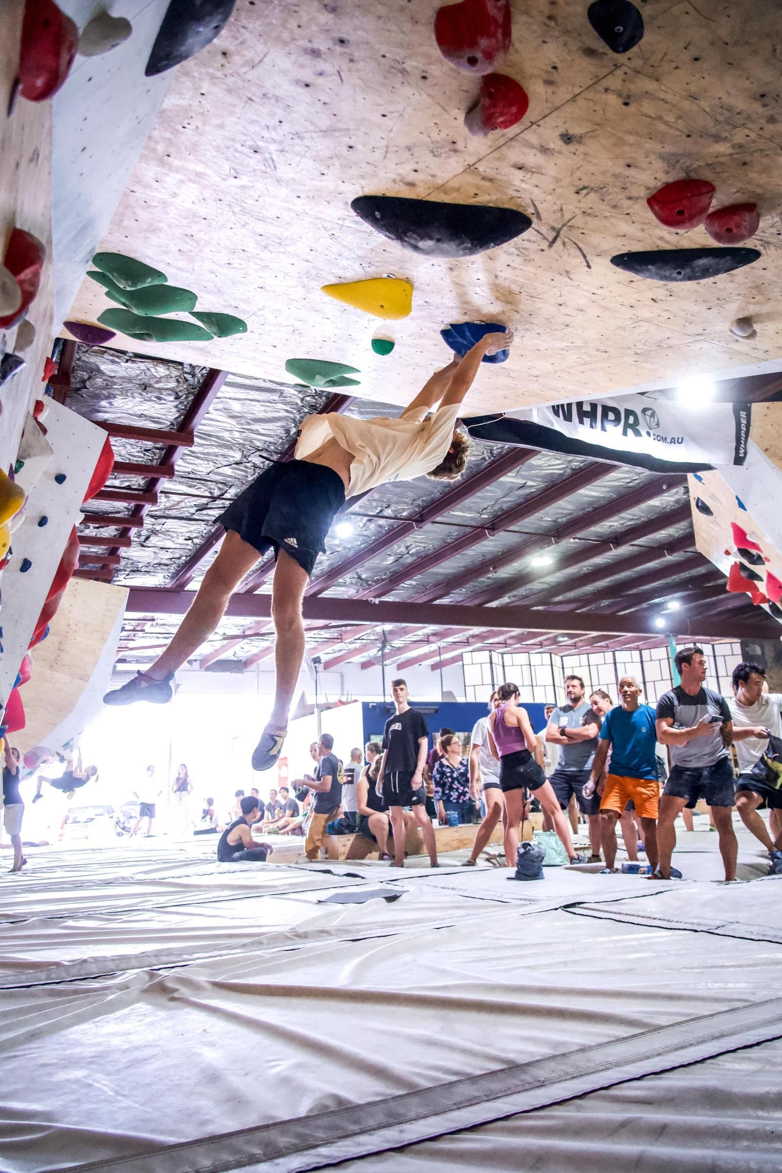 Rock climber in mid-air, reaching for a hold on an indoor climbing wall. Spectators watch below.