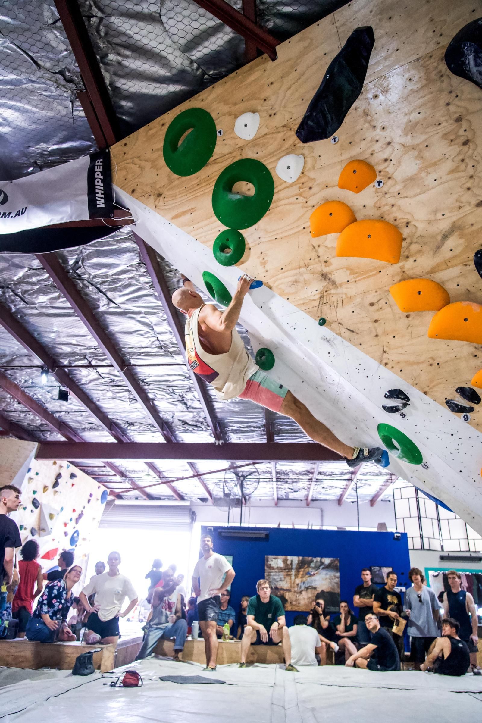 Climber on an indoor wall, reaching for a hold. Spectators watch below. Wall has colorful climbing holds.