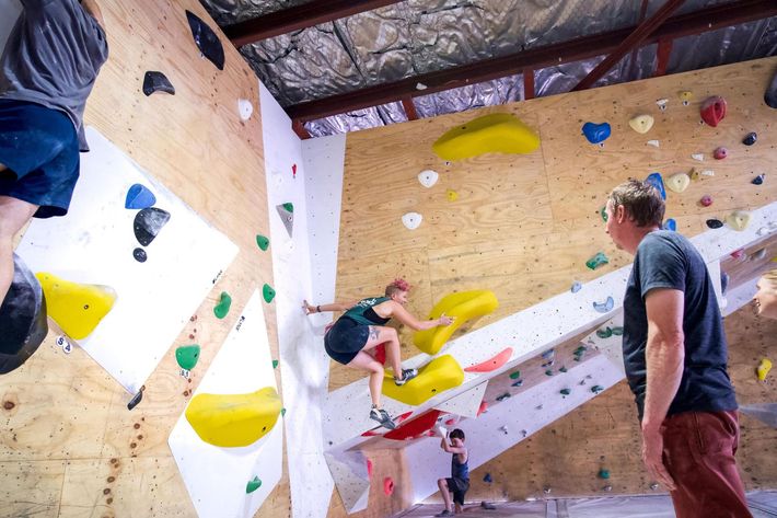 People bouldering indoors; woman on wall with yellow holds, man spotting.