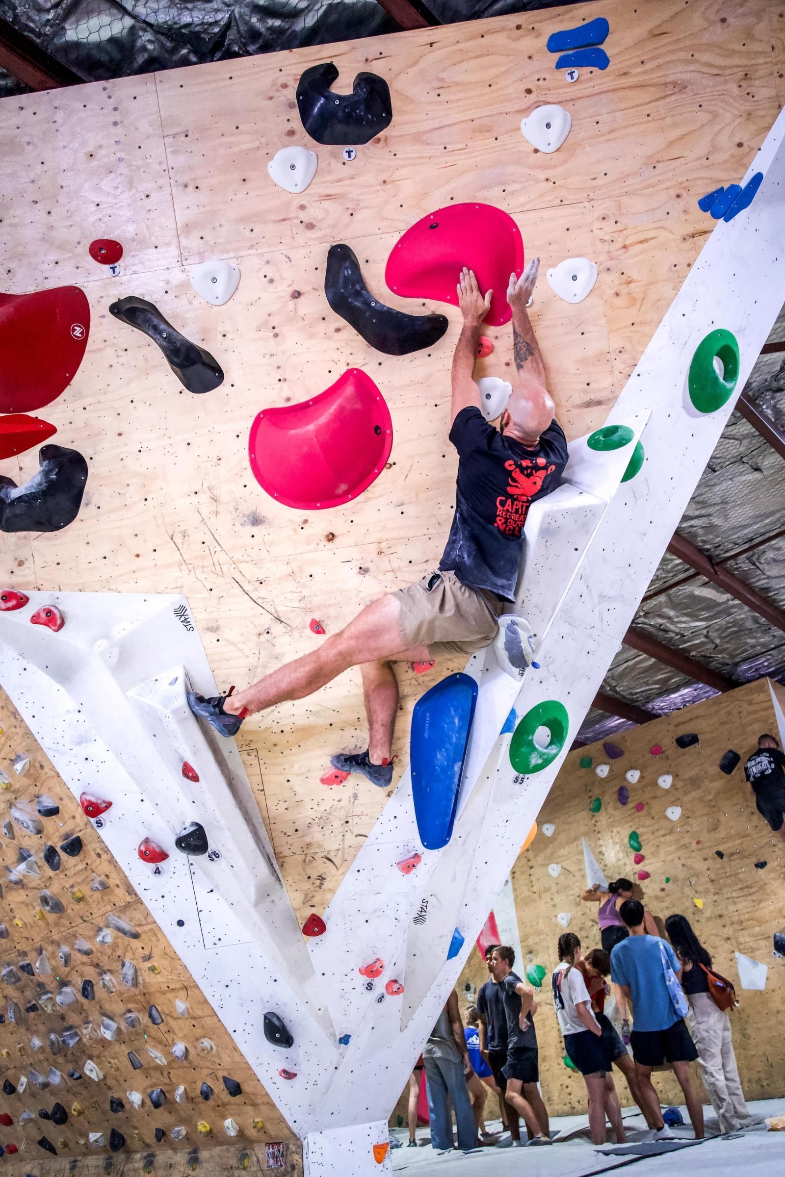 A person bouldering on an indoor climbing wall, reaching for a large pink hold. Other climbers watch.