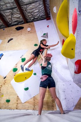 Two people bouldering, one reaching for a hold while the other spots them. Indoor climbing gym.