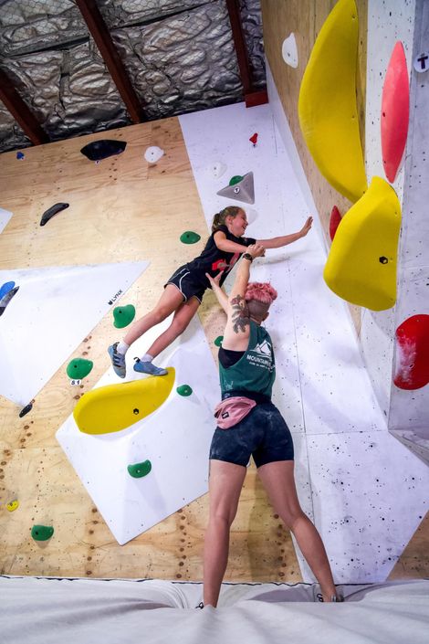 Two people bouldering, one reaching for a hold while the other spots them. Indoor climbing gym.