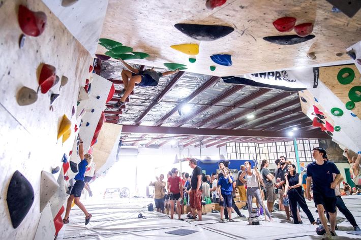 People climbing on indoor rock walls at a climbing gym. Spectators watch.