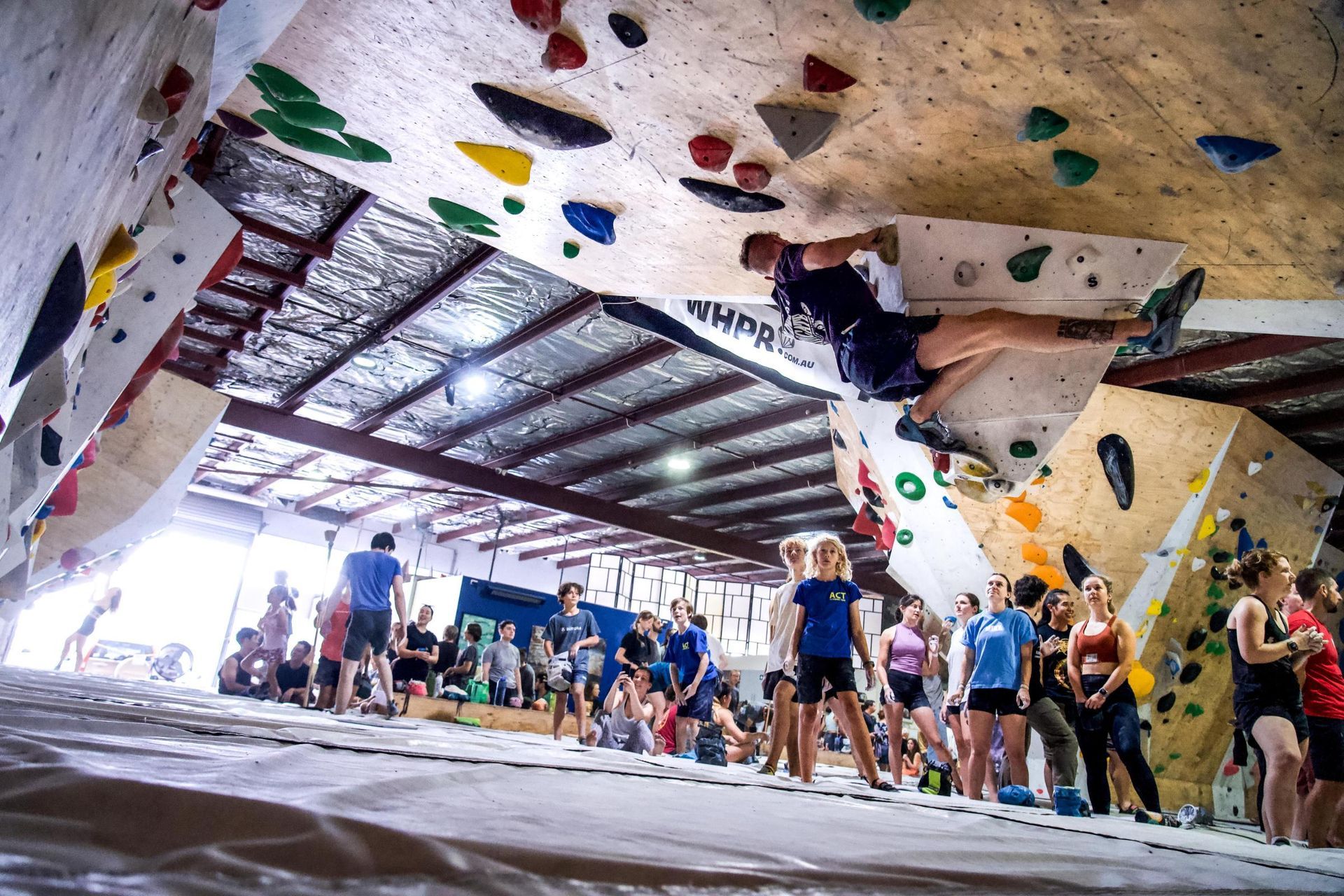 Man bouldering in an indoor climbing gym, above crowd. Colorful holds on walls.