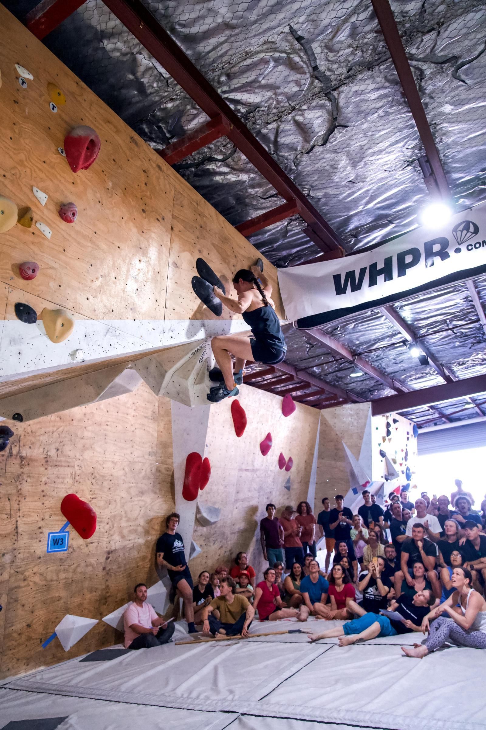Rock climber ascending a wall with colored holds, indoors at a competition. Spectators watch.