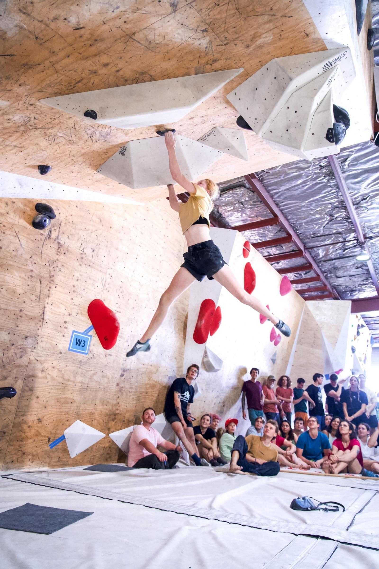 A woman rock climbing, mid-air with legs split. Others watch, inside a climbing gym.