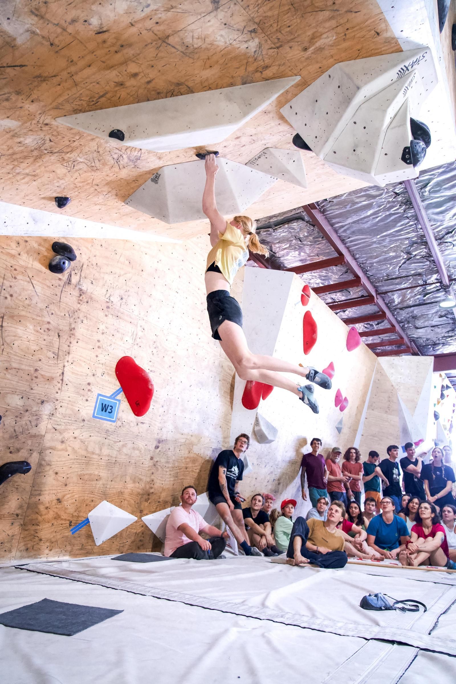 Woman rock climbing in an indoor gym, jumping for a hold, with spectators watching.
