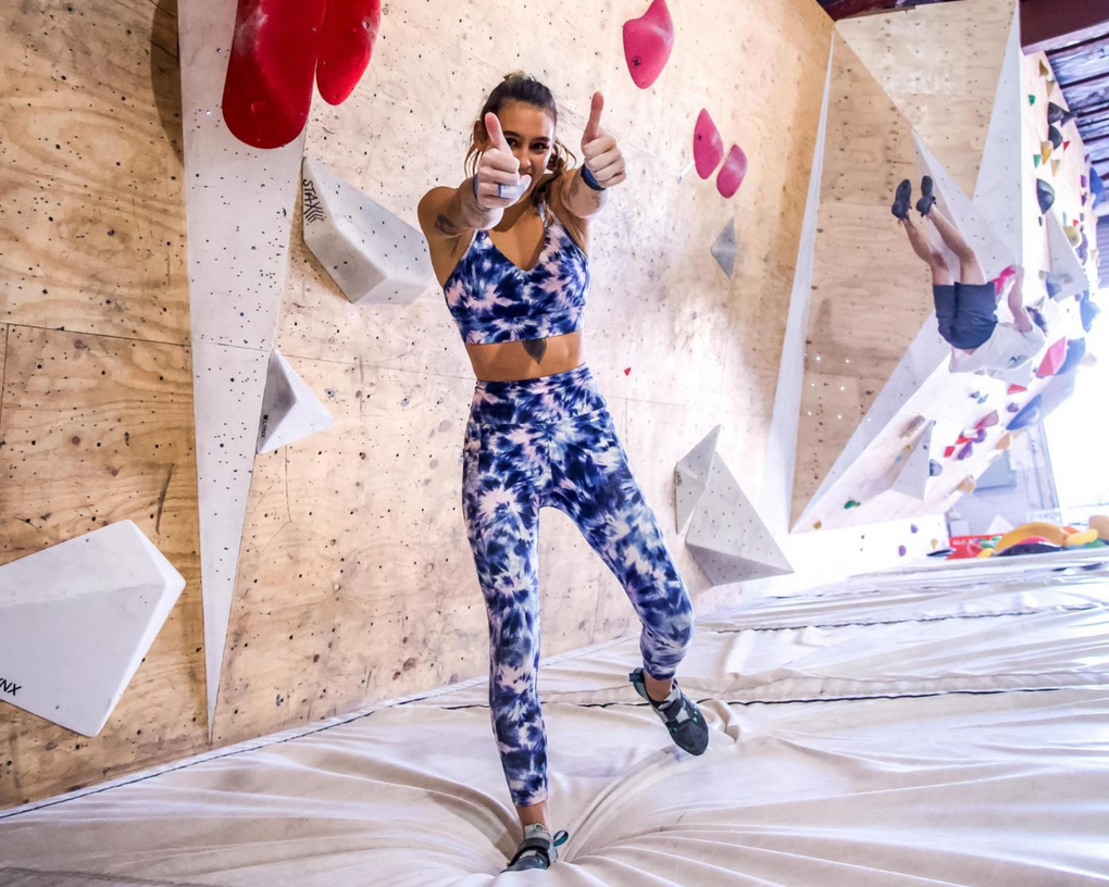Woman in tie-dye activewear gives thumbs up on a climbing gym mat, climbing wall in background.