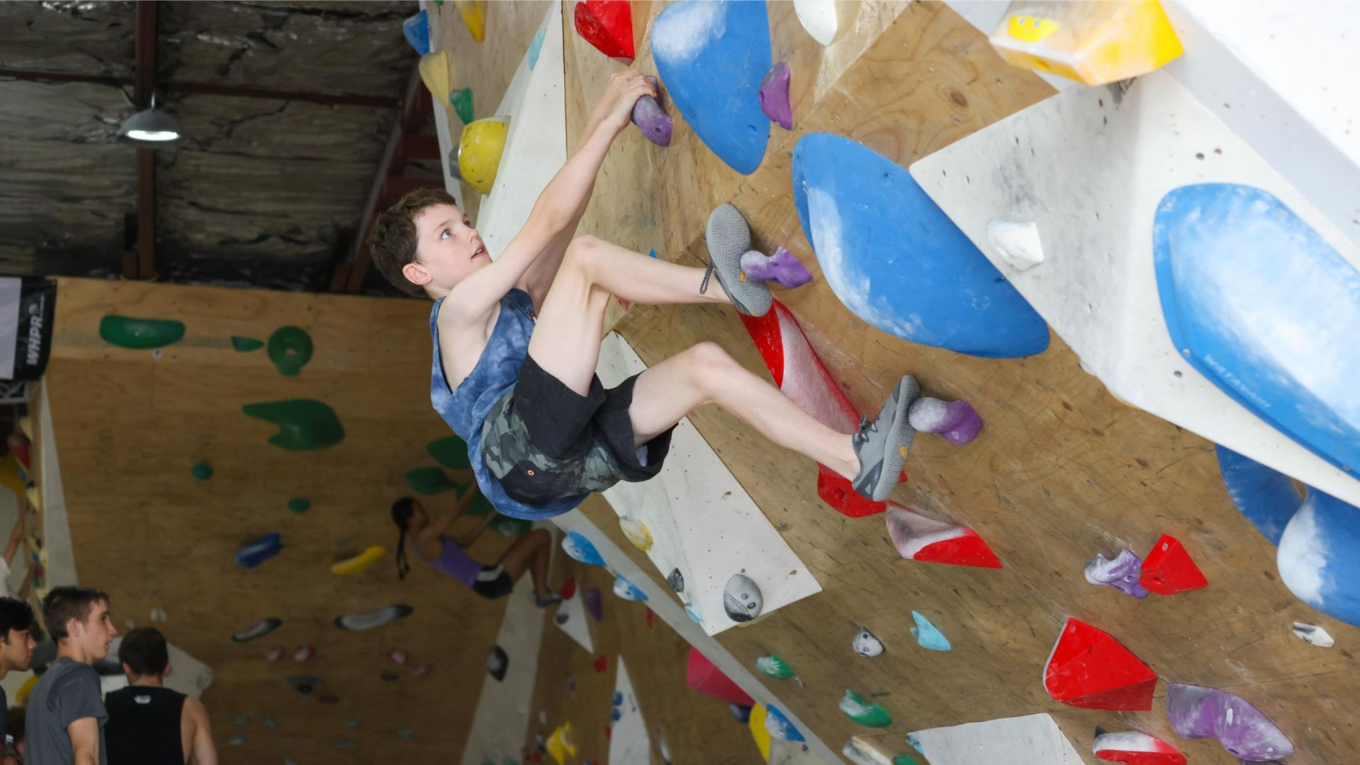 Person climbing a wooden indoor rock wall with blue and white handholds.