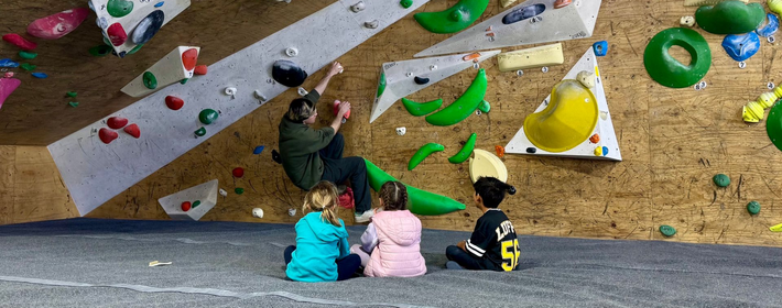Children watching a climber on an indoor bouldering wall with colorful holds