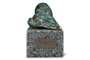 Lille bronzeskulptur på en grå piedestal med indgraverede koordinater.