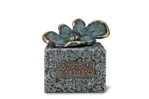 Lille grå stenblok med en blå-og-guld blomsterskulptur på toppen og en rød nummereret plakette på forsiden