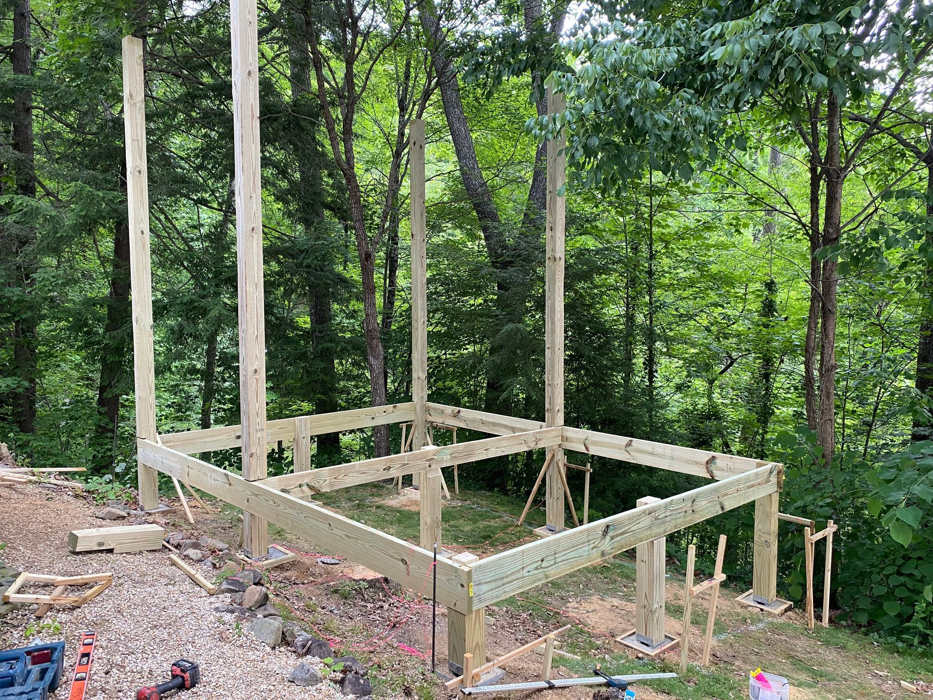 Wooden deck frame under construction in a wooded area, with tall posts and supporting beams.
