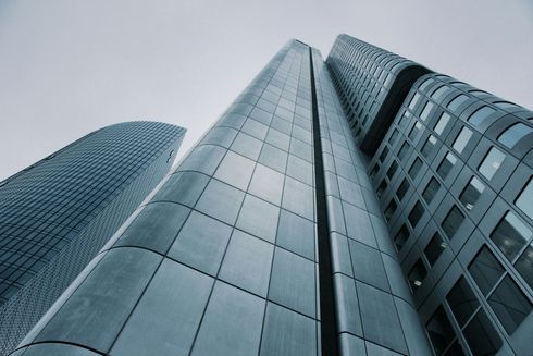 Looking up at modern skyscrapers with gray-blue steel and glass facades against a cloudy sky.