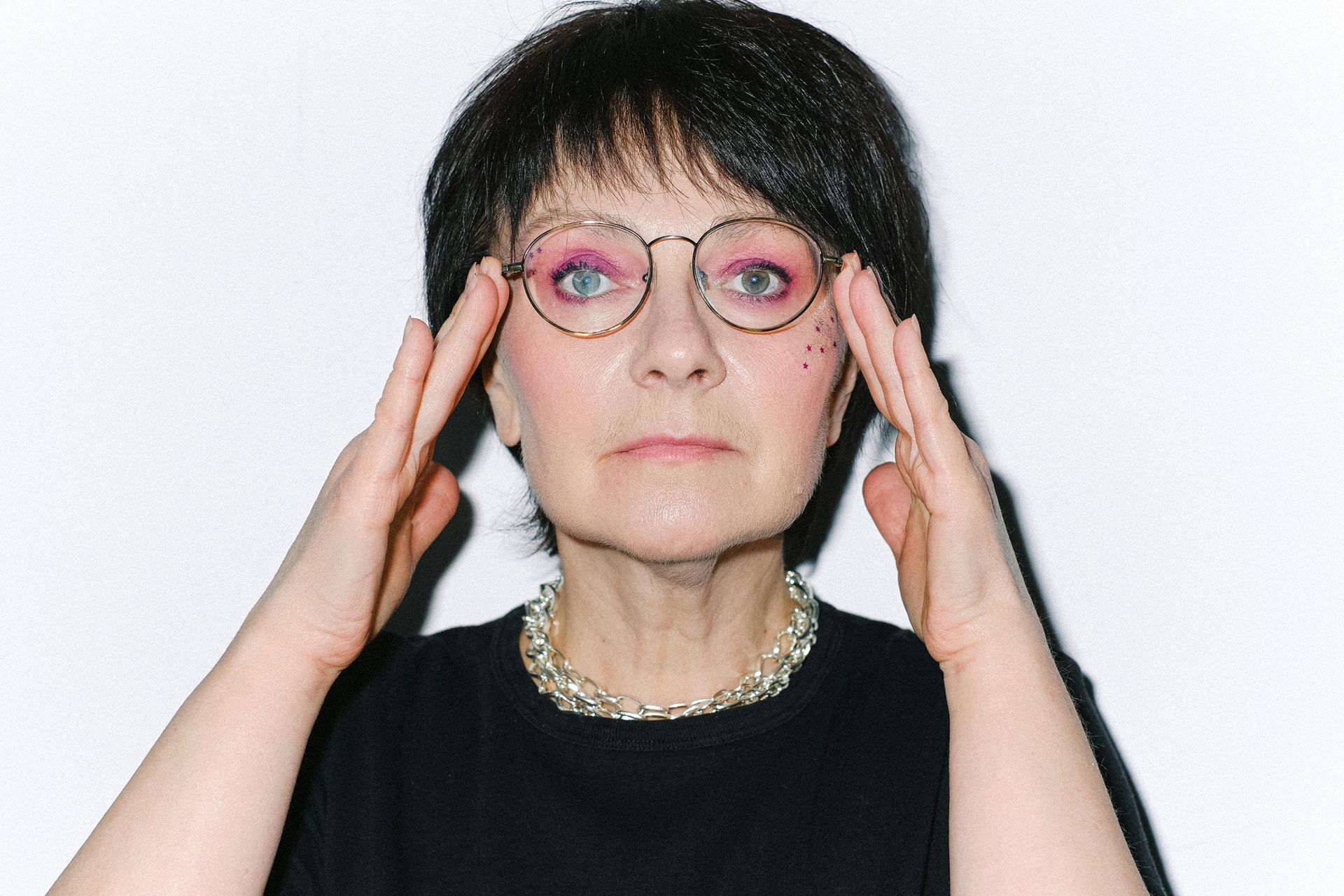 Woman with glasses, hands to head, looks at camera. Dark hair, black top, silver necklace, white background.