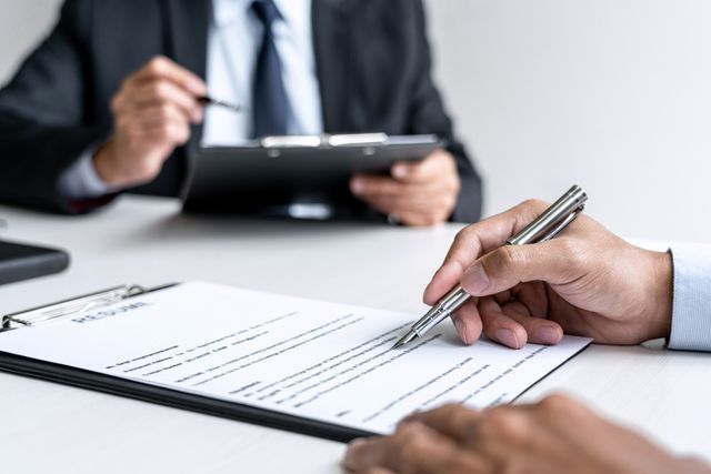 Person signing a document on a clipboard during a meeting. Another person is in the background.