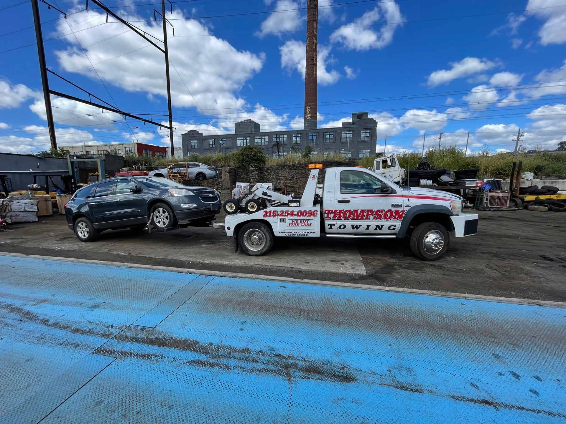A white Thompson Towing truck pulls a gray SUV in a gravel yard under a bright, partly cloudy blue sky.