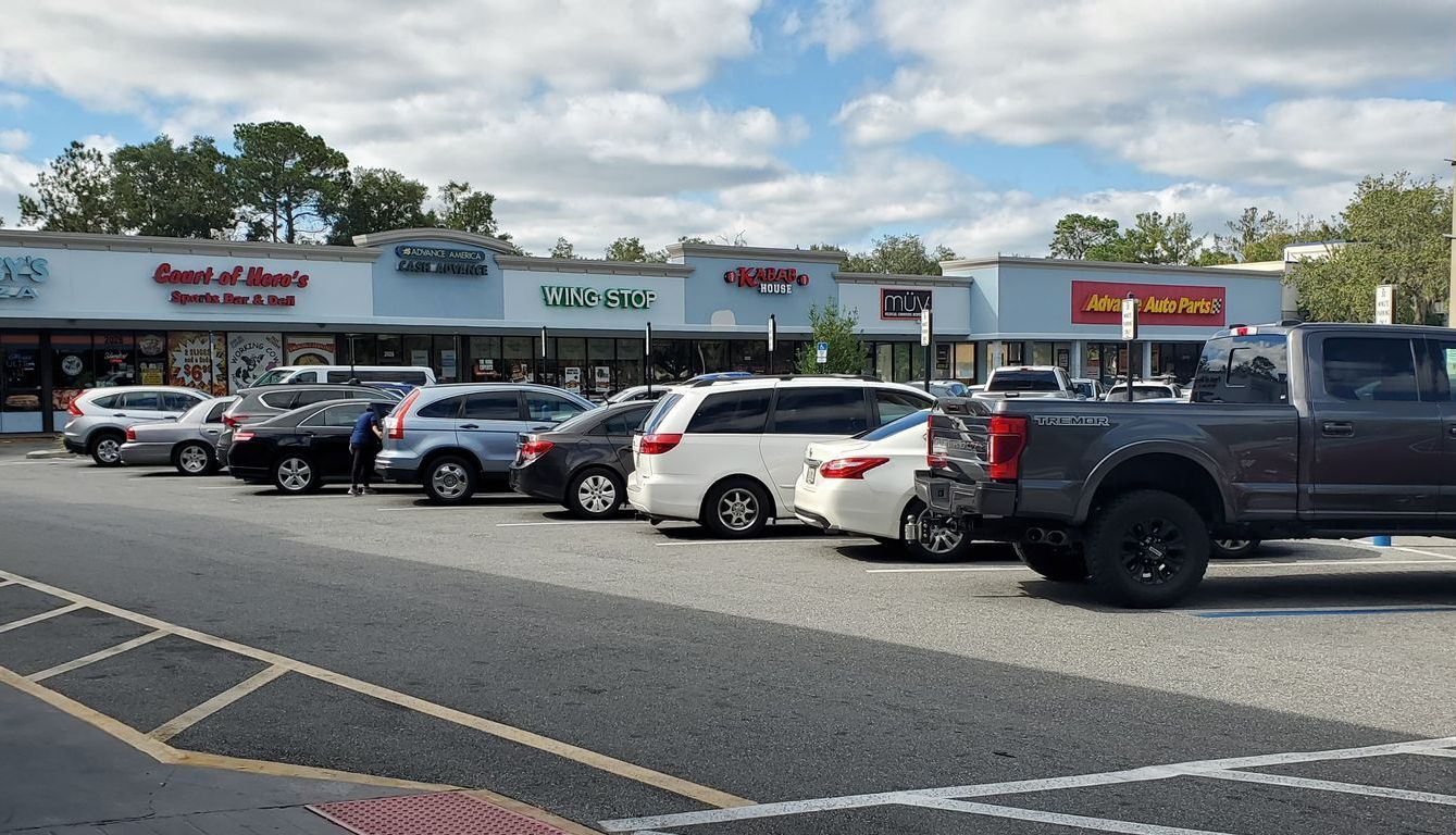 A row of cars are parked in a parking lot in front of a shopping center.