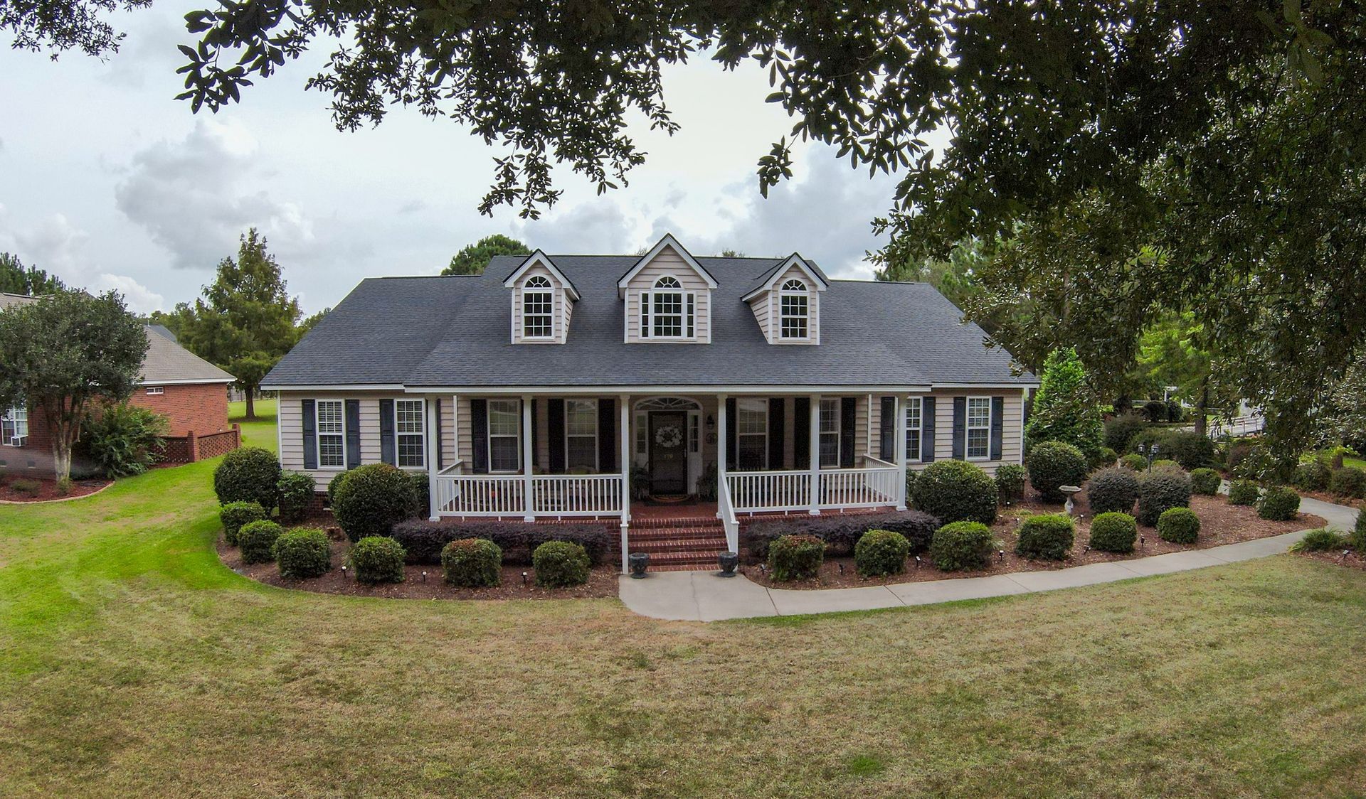 A one-story brick house with a front porch, black shutters, and three dormers under a cloudy sky.