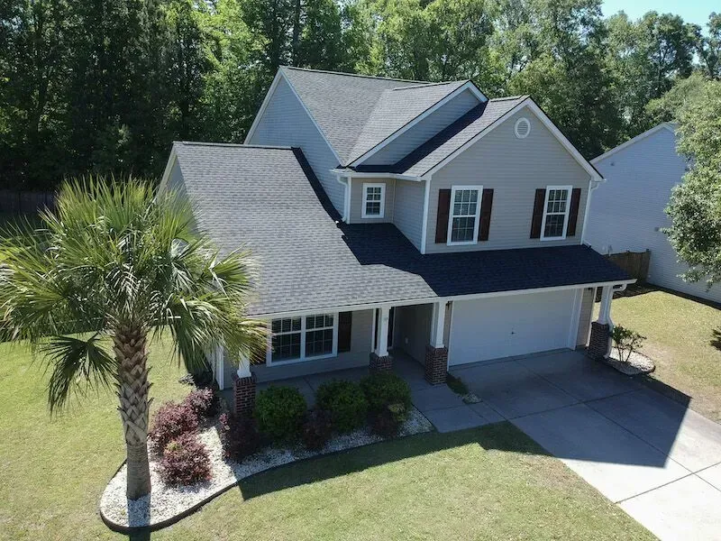 Two-story beige house with gray roof, brown shutters, and palm tree in front yard.
