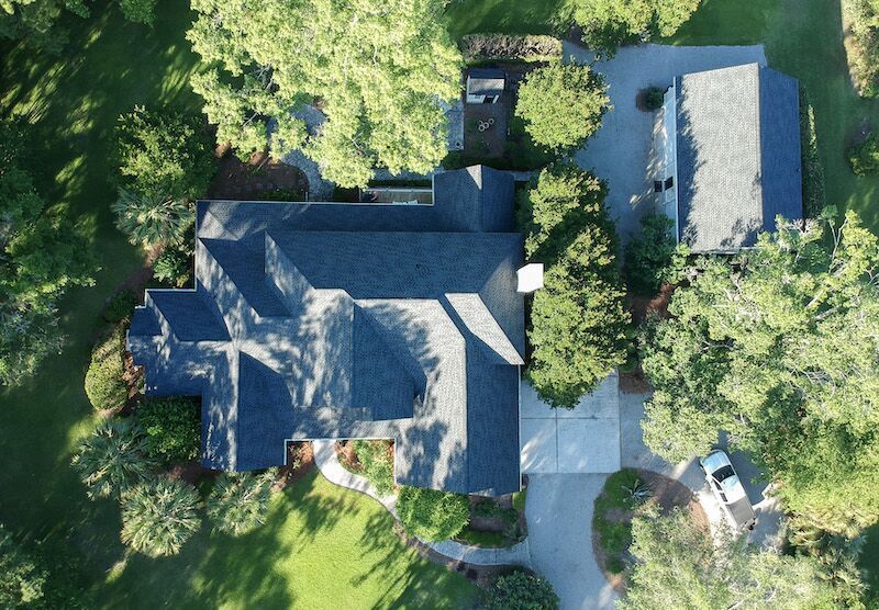 Aerial view of a house with a dark roof and a driveway, surrounded by trees and greenery.