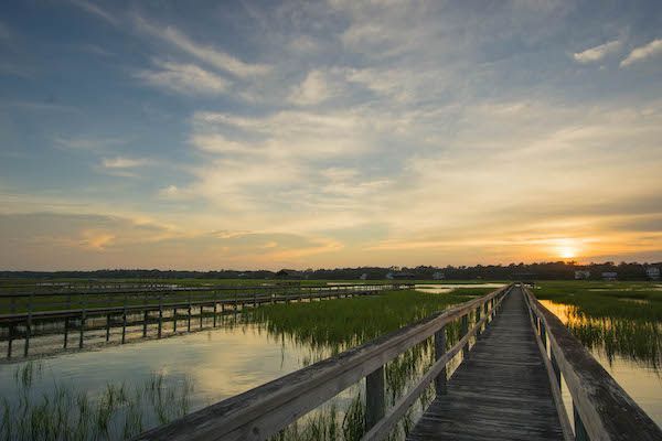 Wooden boardwalk over marsh at sunset; golden sky reflects in water.