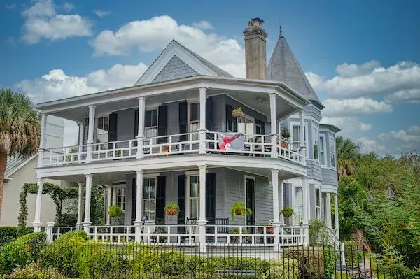 Two-story gray Victorian house with wraparound porch, white railing, and a small tower under a partly cloudy sky.