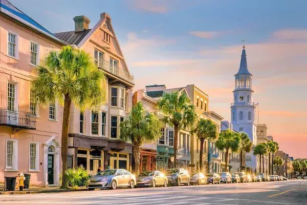 Historic Charleston street with colorful buildings, palm trees, and a steeple at sunset.