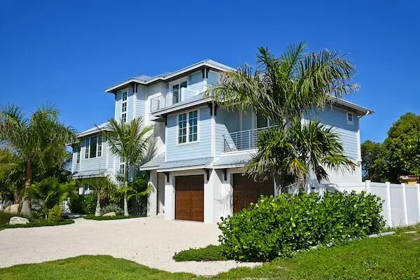 Two-story blue house with brown garage doors, palm trees, and clear blue sky.