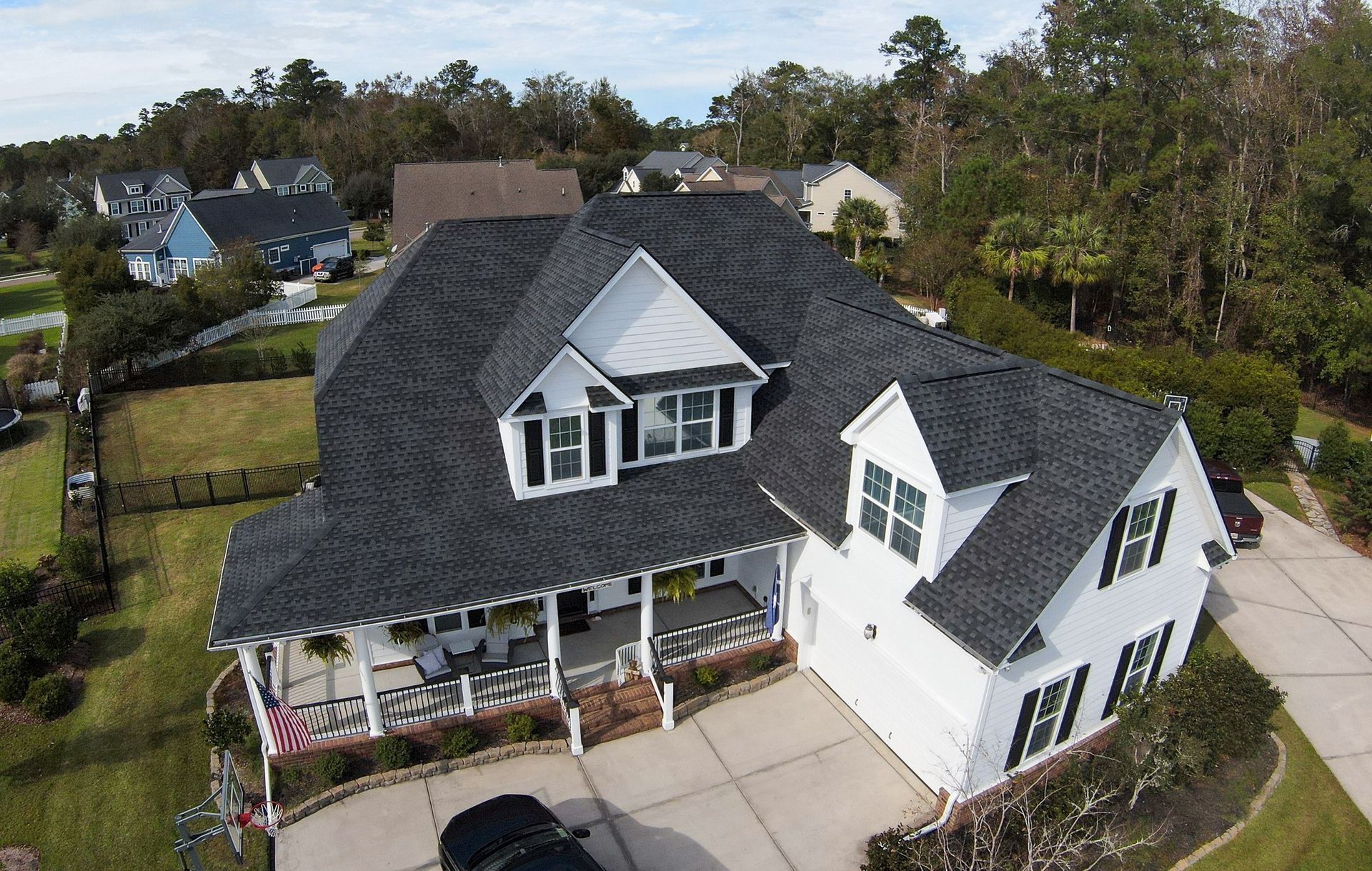 White house with a dark gray roof and black shutters, porch with seating, green lawn, trees, and blue sky.