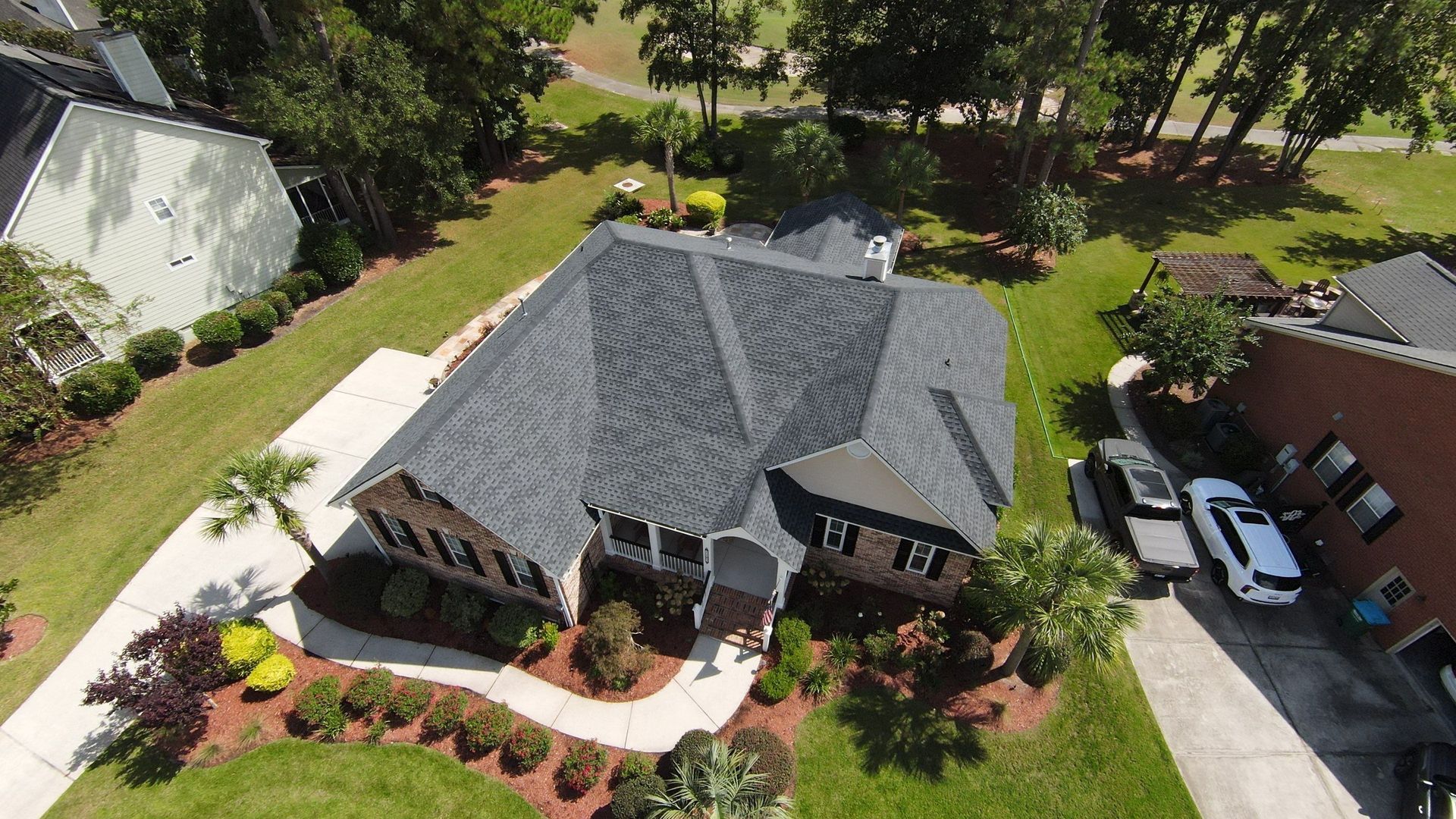 Aerial view of a house with a dark gray roof, surrounded by green grass and landscaping, with a driveway and cars.