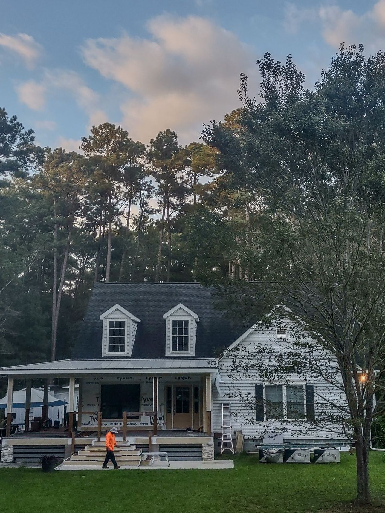 House under construction with worker, porch, dormer windows, and trees in background. Cloudy sky.