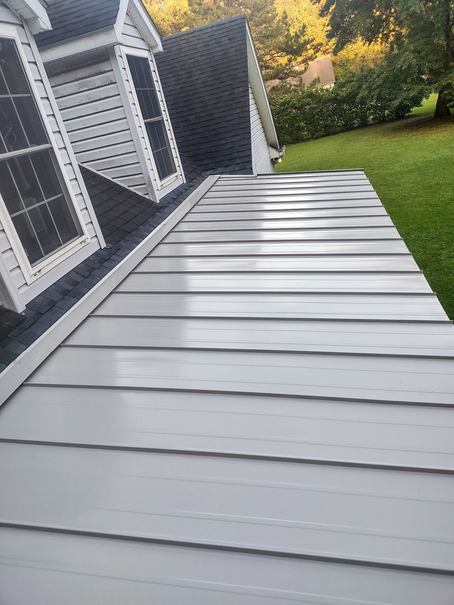 Gray metal roof on a house, with black roofing and white trim, overlooking green grass and trees.