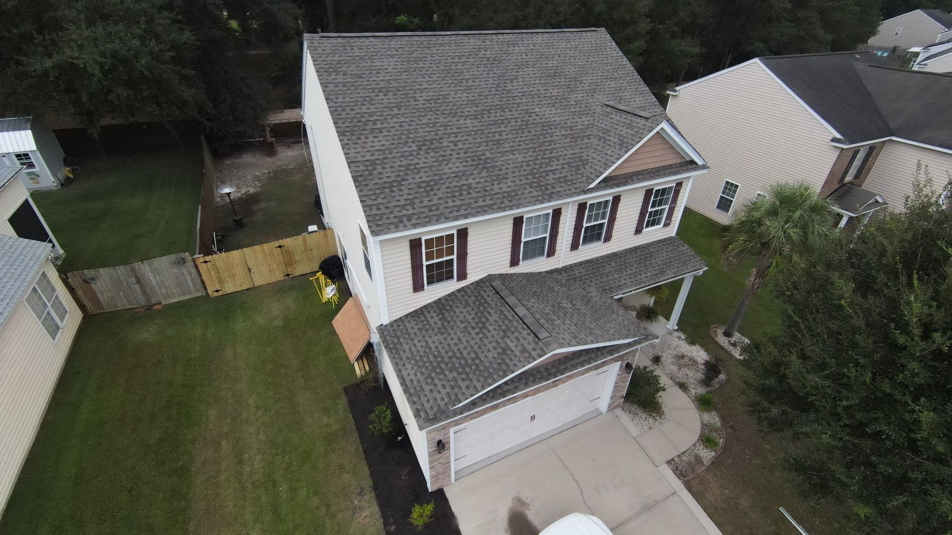 Aerial view of a two-story beige house with gray shingle roof, surrounded by green grass and trees.