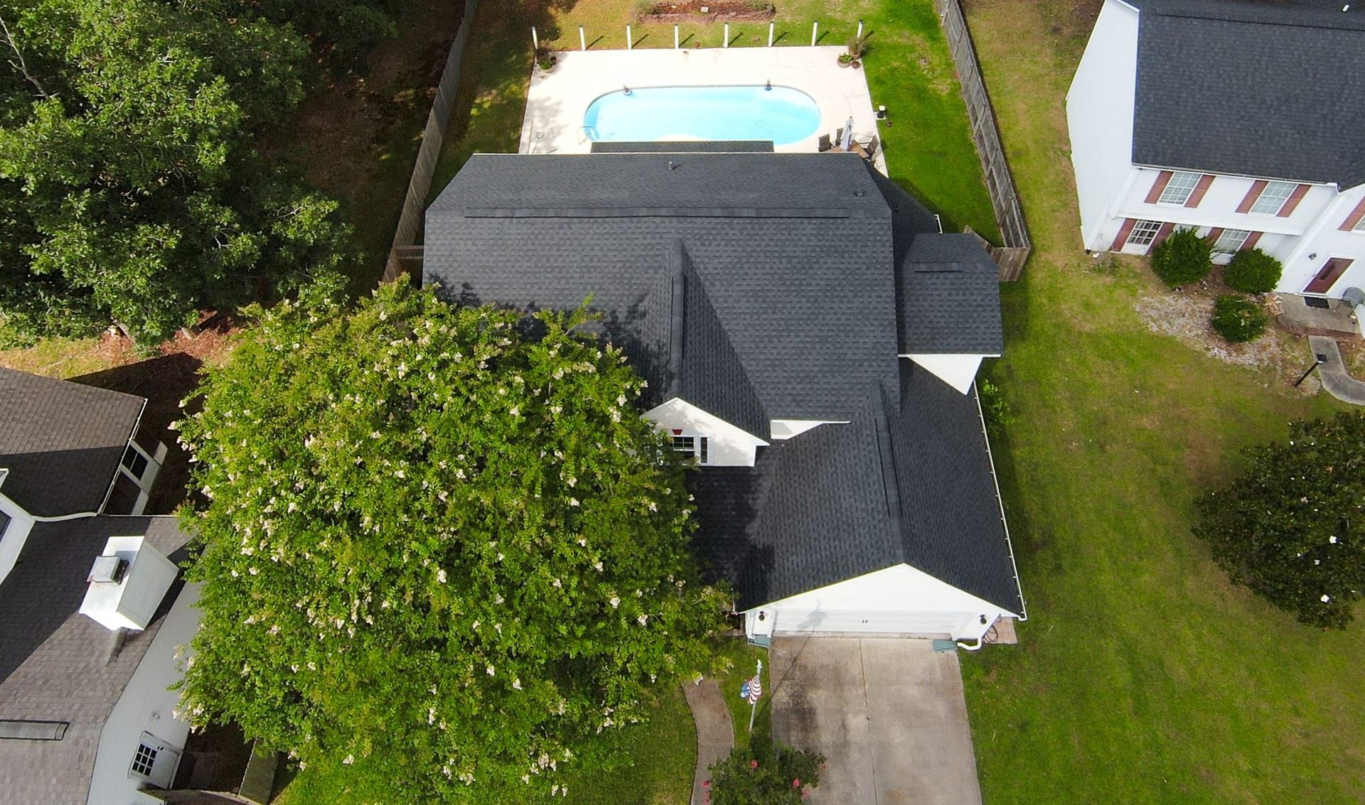 Aerial view of a house with a black roof, a driveway, and a backyard pool surrounded by green grass and trees.