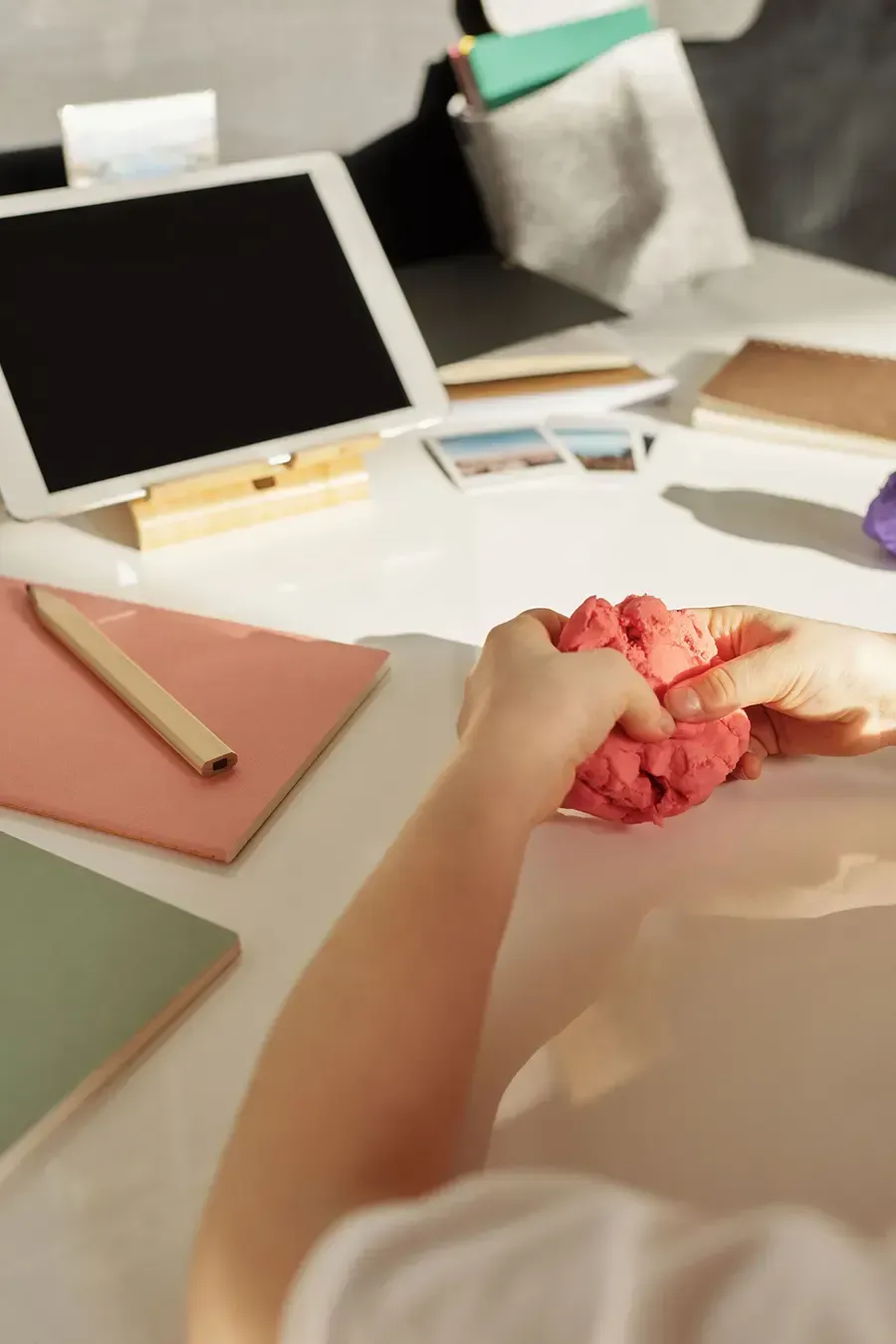 Person kneading pink clay at a cluttered desk with a small blank monitor and notebooks