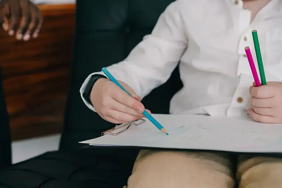 Person writing on a clipboard with a blue pencil while holding colorful markers in a white shirt