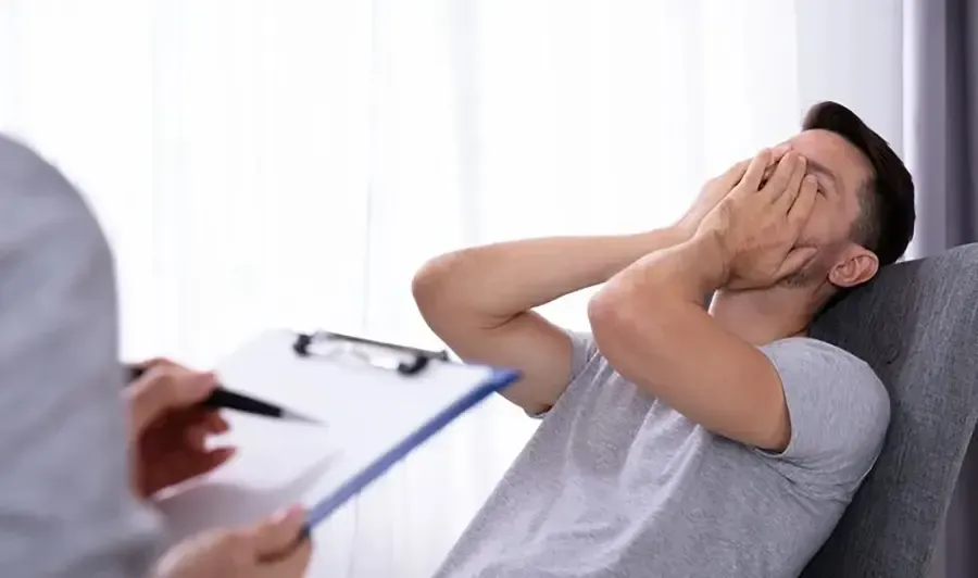 A man leaning back in a chair while holding his hands over his face as he talks to a therapist