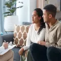 Two people listening to a therapist in a bright living room, seated on a couch.