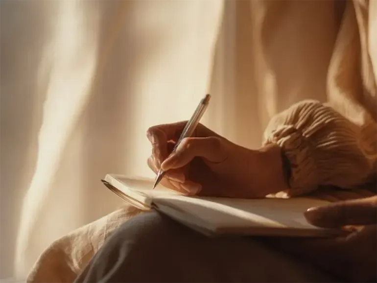 Hand writing in a notebook beside a warm-lit window
