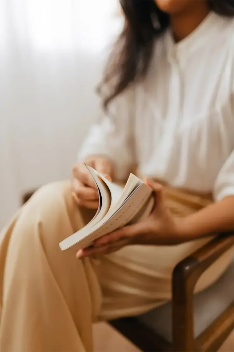 Person holding an open book while seated in a wooden chair, wearing a white top and tan pants.