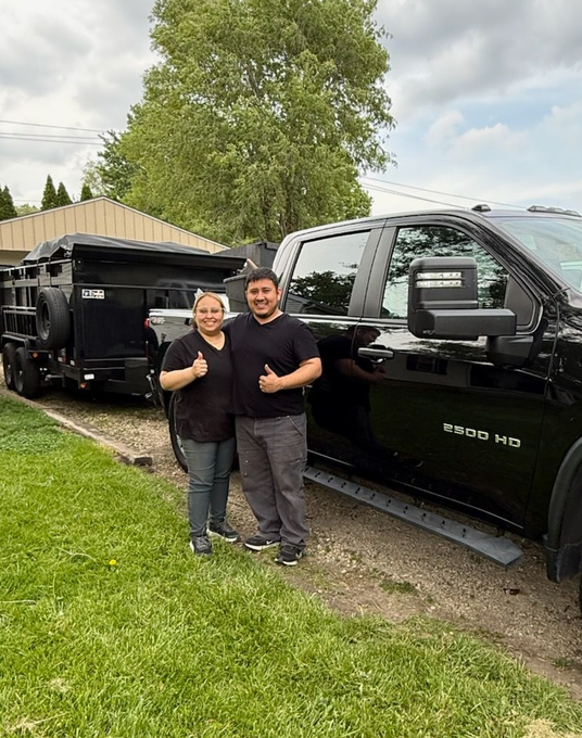 Two people standing beside a black HD pickup truck and trailer on a grassy roadside, smiling and giving thumbs up