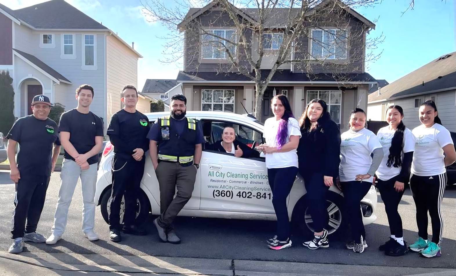 Group of people posing beside a white car in a suburban driveway under a sunny sky