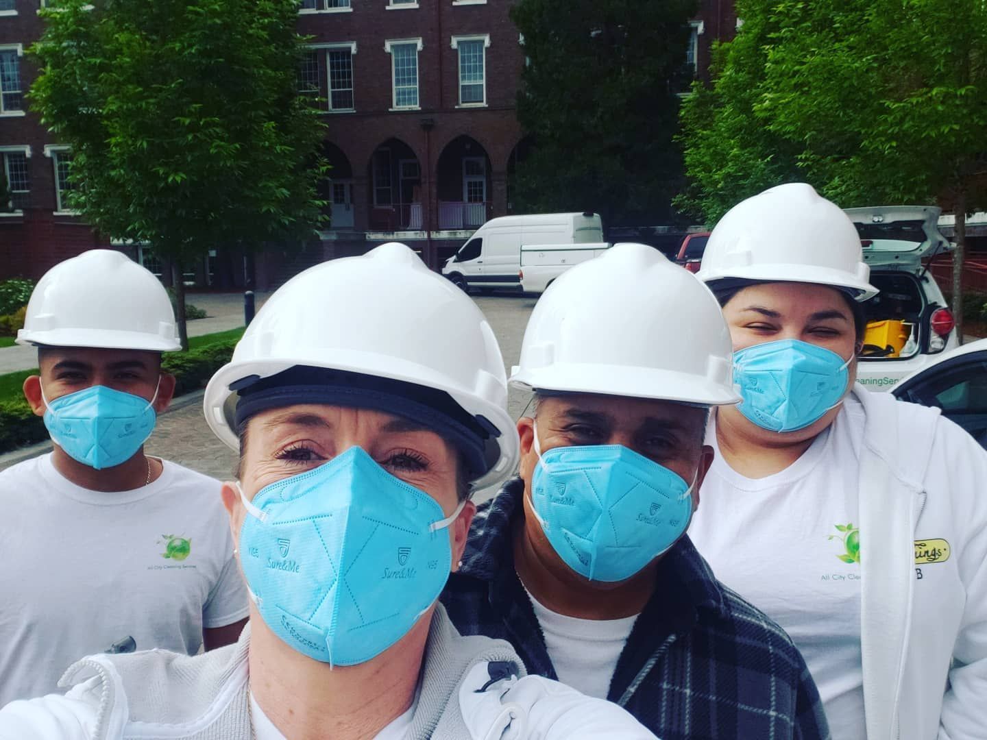 Four workers in hard hats and blue masks standing outdoors in front of a brick building.
