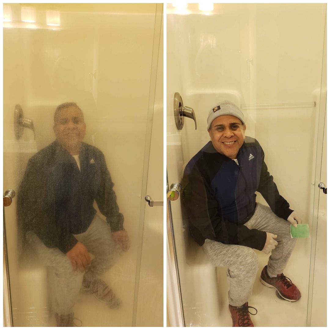 Two men sitting in a shower stall, one blurred and one smiling, with a yellowish bathroom light.