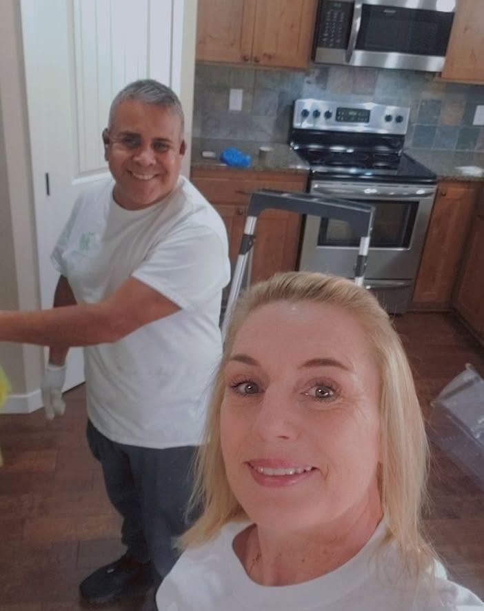 Two people smiling in a kitchen with wooden cabinets and a stainless steel stove.