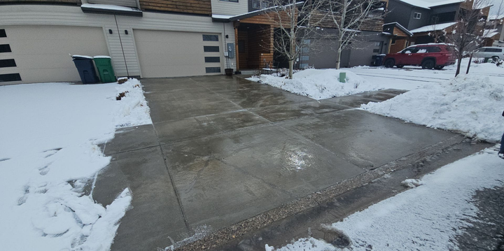 A concrete driveway and sidewalk area cleared of snow in front of a house with two garage doors and a red car parked nearby.